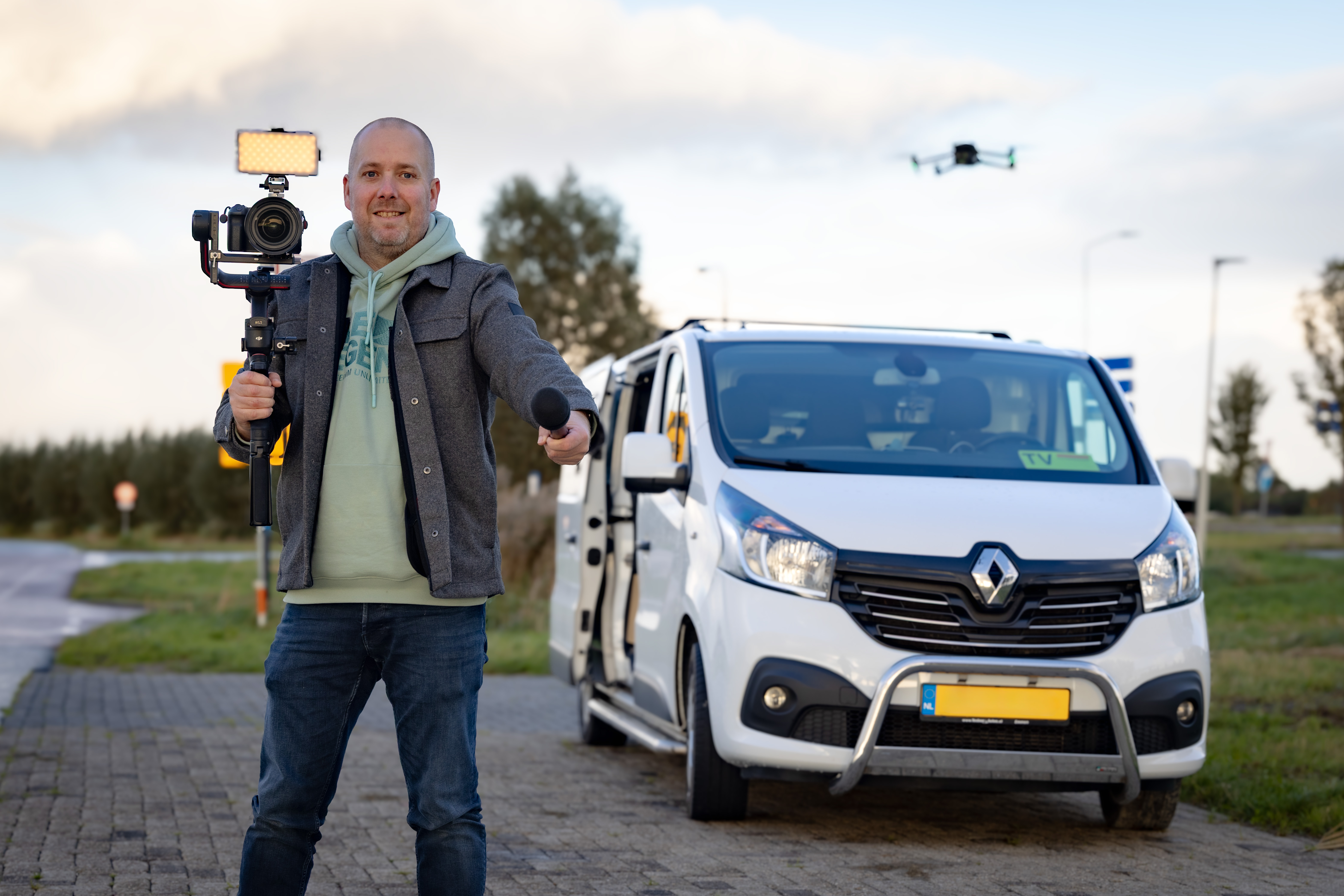Jaring Rispens met camera en microfoon naast zijn werkbus, met een drone op de achtergrond